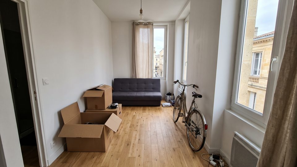 Petit studio vu depuis la porte, cartons de déménagement entrouverts et vélo contre le mur, fenêtre sur une façade en pierre blonde